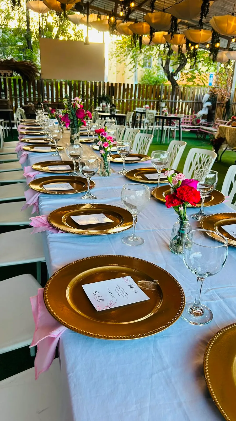 Mesa larga con vajilla dorada, servilletas rosadas y flores.